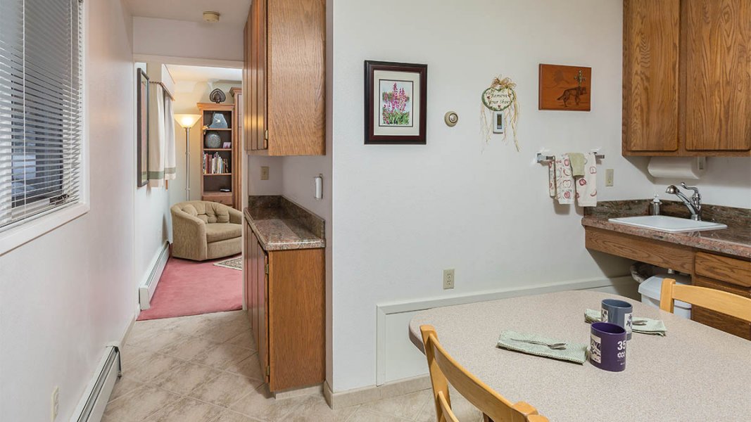 Kitchette with table with chairs, sink, pictures on walls, hallway with cabinets both sides, salmon carpeted sitting room.