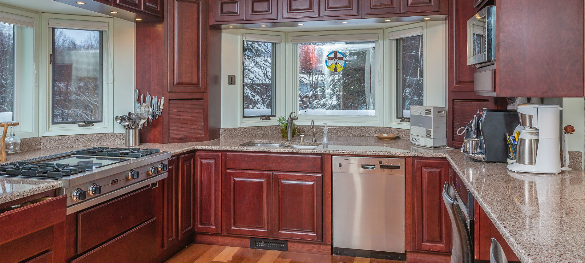 Kitchen with stainless steel appliances and cherry cabinets on all sides, bay windows, marble counters, wood floor.