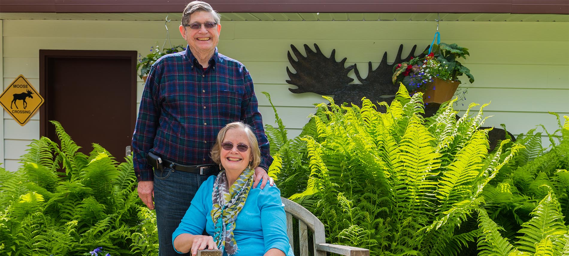 Craig standing next to Caroline seated in garden with lush green ferns and moose silhouette in background