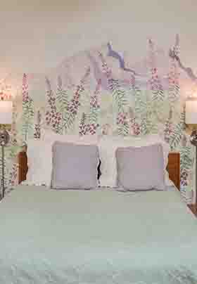 Wall scene above queen bed shows fireweed blooms on purple mountain peaks; wall lamps on each side of bed