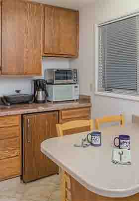 Wall of walnut kitchen cabinets; white sink, microwave, refrigerator, toaster oven, coffee maker; counter with four chairs