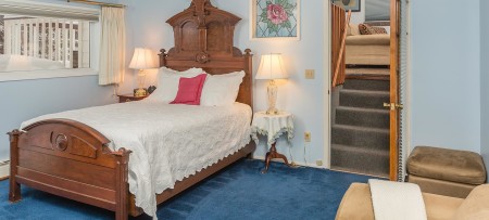 Antique queen bed with white spread; beige chair and ottoman; deep blue carpet; stairs leading to upper room with sofa