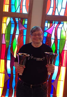 Craig in black F flat=mc squared shirt.; holding two handbells; stained glass window background