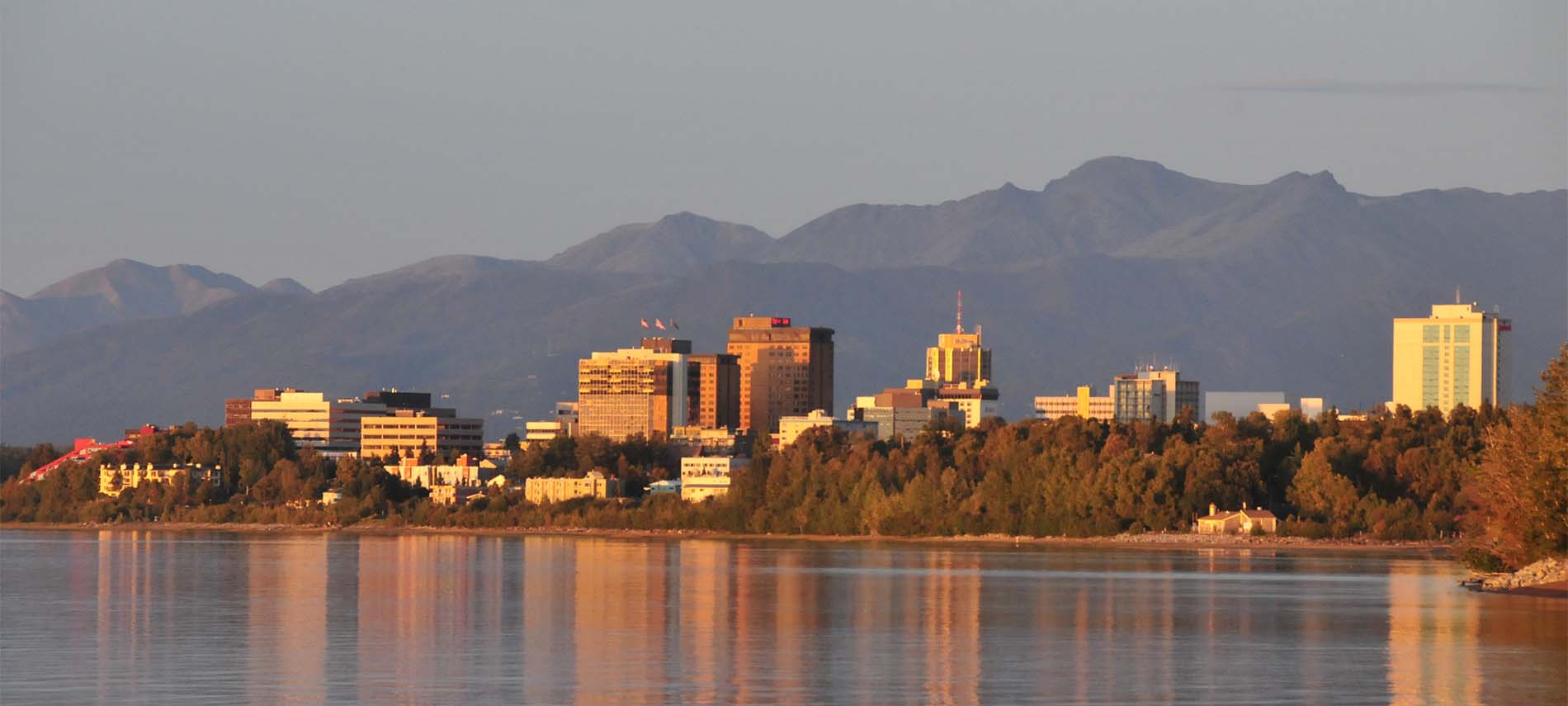 Snow-free mountain background with sun-drenched tall city buildings reflected in foreground’s lake; fall’s orange tinged trees surround lake