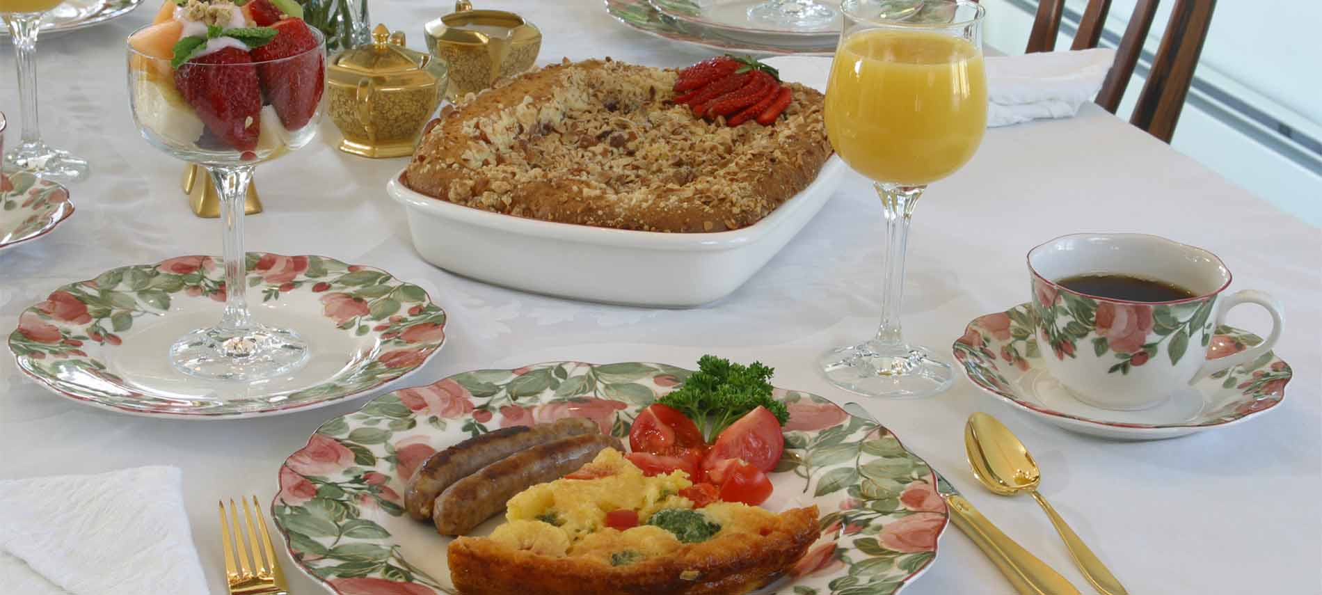 Quiche slice with sausages, tomato, parsley on plate with rose, green foliage pattern; cup of coffee; colorful fruits in goblet