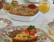 Quiche slice with sausages, tomato, parsley on plate with rose, green foliage pattern; cup of coffee; colorful fruits in goblet