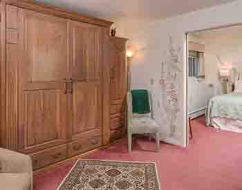 Queen bed with mint green spread, Wooden armoire, white chairs, salmon colored carpet with flowered rug.