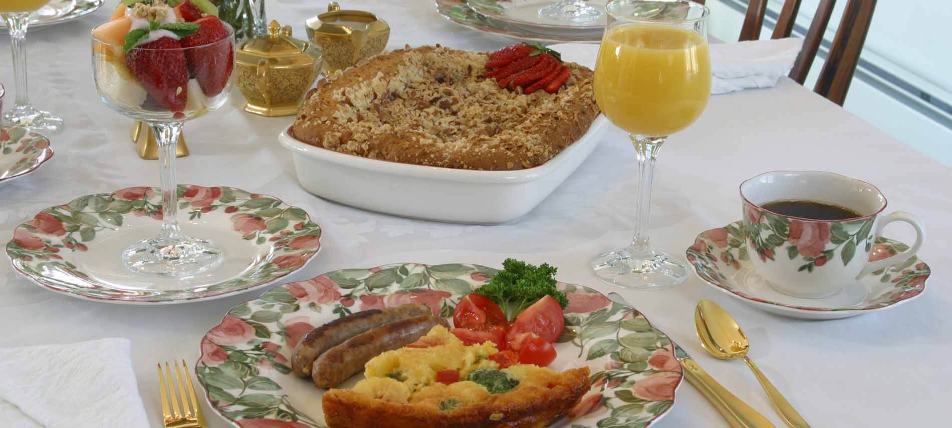 Quiche slice with sausages, tomato, parsley on plate with rose, green foliage pattern; cup of coffee; colorful fruits in goblet.