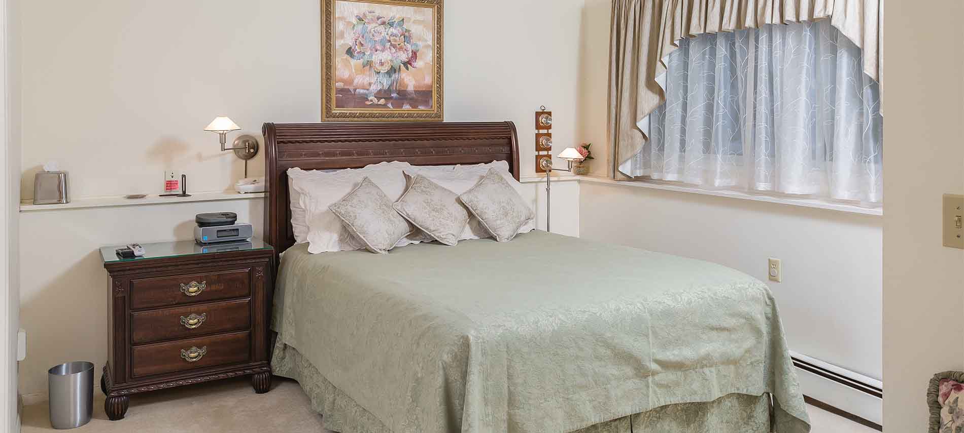 Queen bed with green damask spread; green cushioned bench in foreground; framed pink flower picture above bed; light beige carpet