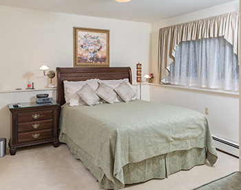 Queen bed with green damask spread; green cushioned bench in foreground; framed pink flower picture above bed; light beige carpet