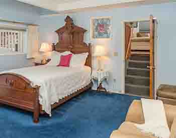 Antique queen bed with white spread; beige chair and ottoman; deep blue carpet; stairs leading to upper room with sofa