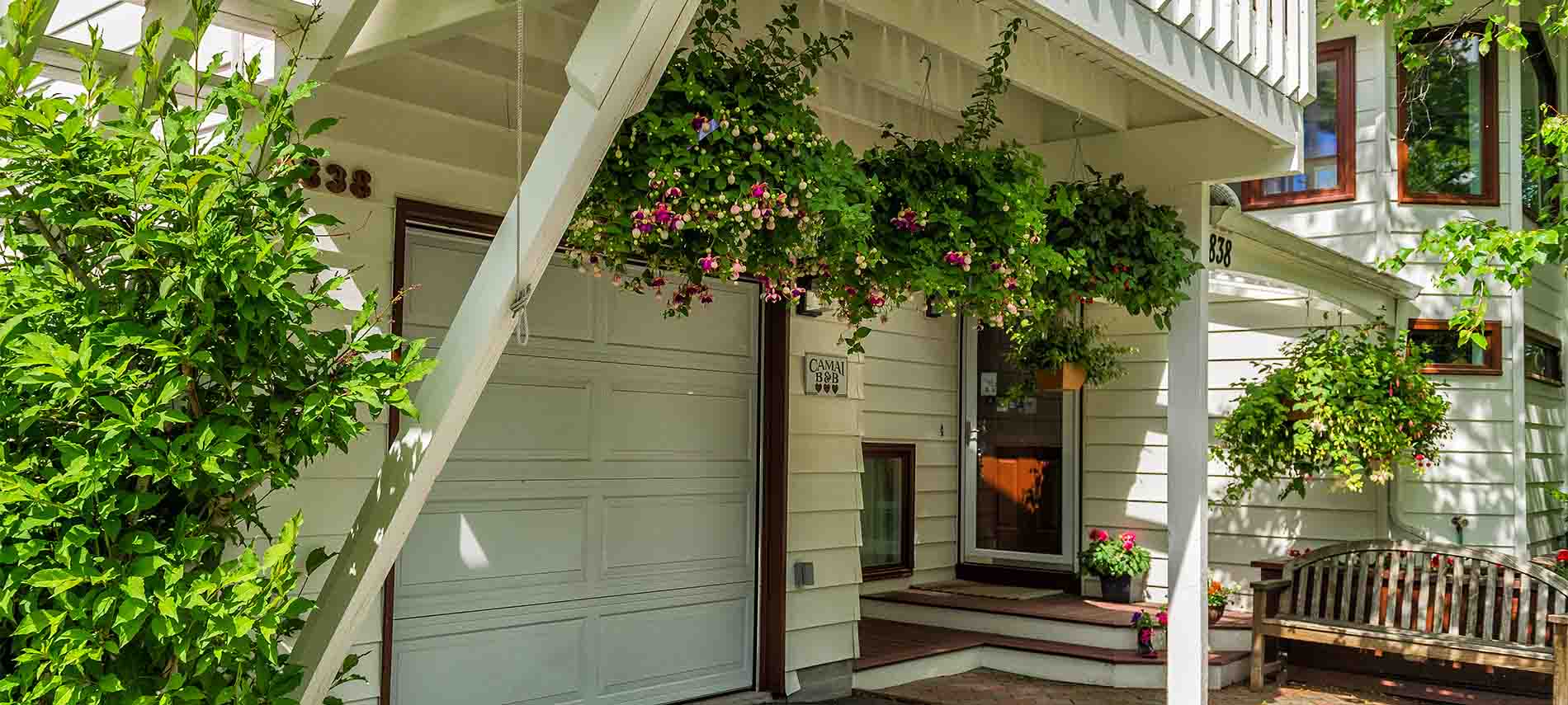 White upper deck with hanging pink fuchsia hanging baskets leads to front entry’s deck with garden bench